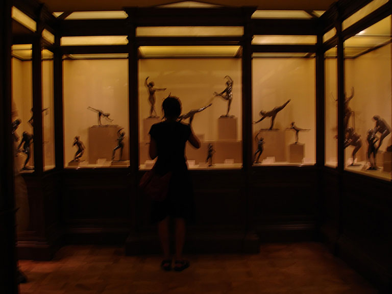 Standing at the lit vitrines of Degas bronzes — dancers mid-leap on their plinths — sketchbook raised, silhouetted in the amber museum light