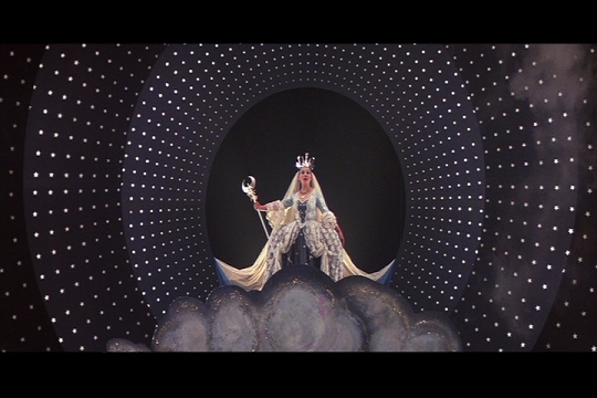 Film still of the Queen of the Night: a crowned soprano with a crescent-moon staff rises from painted clouds inside a swirling tunnel of stars